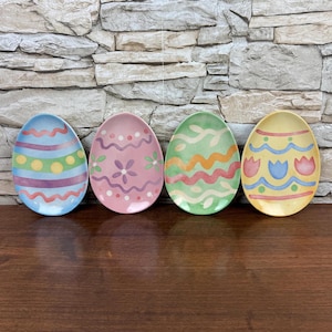 May include: Four decorative Easter egg-shaped plates. Each plate is painted with a unique design in pastel colors. The designs include stripes, dots, flowers, and wavy lines. The plates are arranged on a wooden surface against a stone wall.