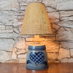 May include: A table lamp with a cylindrical blue ceramic base featuring a dark blue cross-hatched heart design. The lamp is topped with a conical, textured tan fabric lampshade that emits a warm glow from the light source inside the base.