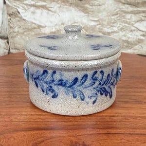 May include: A small, round, lidded ceramic jar with a light gray base and a blue floral design. The jar has a small knob on top and two small handles. The lid features a matching blue floral pattern.