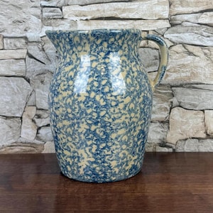 May include: A blue and cream-colored ceramic pitcher with a speckled pattern. The pitcher has a handle and a spout for pouring. The background is a stone wall. This pitcher is suitable for serving beverages.