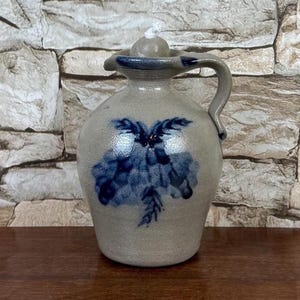 May include: A stoneware jug with a handle and a lid. The jug is light gray with a blue floral design. The jug is sitting on a wooden surface.