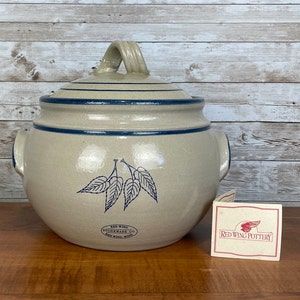 May include: A beige stoneware crock with a blue trim and a leaf design. The crock has a lid with a handle. The crock is labeled "Red Wing Stoneware Co. Red Wing, Minn."