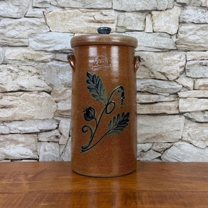 May include: A brown ceramic crock with a lid and handles. The crock features a floral design in black and green. The lid has a small, dark knob. The crock is cylindrical and sits on a wooden surface.
