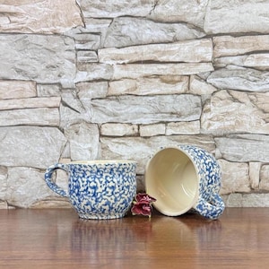 May include: Two ceramic mugs featuring a blue and white speckled pattern on the exterior and a solid cream-colored interior. One mug is positioned upright with a handle on the left, while the other is tilted on its side, revealing the interior.
