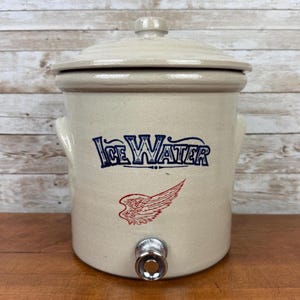 May include: A beige ceramic ice water cooler with a lid and a spigot. The words "Ice Water" are printed in blue, and a red wing graphic is below. The cooler has two handles and sits on a wooden surface.