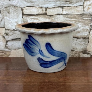 May include: A beige ceramic planter with a scalloped rim and a hand-painted blue tulip design on the front. The pot has a wide opening, a tapered body, and a dark interior. The exterior surface features a speckled texture throughout the clay body.