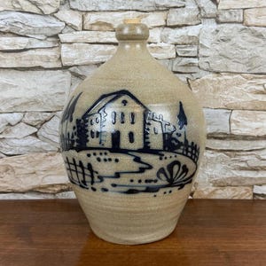 May include: A ceramic bottle with a tan glaze and a black and white illustration of a house and trees. The bottle has a cork stopper.