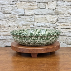 May include: A round ceramic bowl featuring a speckled green and white pattern. The bowl is positioned on a circular wooden stand with short legs. The bowl has a wide, shallow basin with a smooth, rounded rim, showcasing a consistent mottled finish.