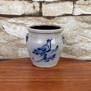 May include: A small, speckled light gray stoneware crock featuring a hand-painted blue bird design on the front. The cylindrical vessel has a thick rim, a dark interior, and two small, circular side handles integrated into the upper body.