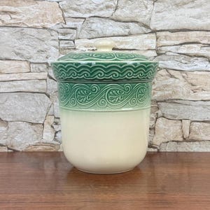 May include: A ceramic canister with a cream-colored base and a removable lid. The upper portion of the canister and the lid feature a raised, embossed green pattern consisting of repeating wave-like scrolls and leaf-like motifs.