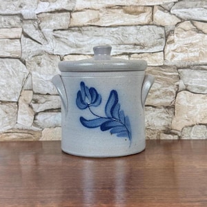 May include: A light gray stoneware crock with a matching lid and two side handles. The exterior features a hand-painted blue floral design with a single flower and leaves. The ceramic surface has a speckled texture throughout the entire body.