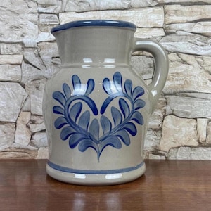 May include: A ceramic pitcher with a light gray body and a blue rim and handle. The pitcher features a blue floral design on the front. The pitcher is sitting on a wooden surface.