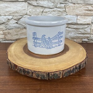 May include: A white ceramic crock with a blue and white design of a wheel and fence. The crock is sitting on a wooden slice.