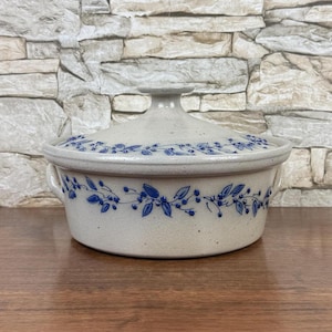 May include: A round, speckled off-white ceramic casserole dish with a matching lid. The dish features a hand-painted blue vine pattern with small leaves and berries encircling the exterior and the lid. It includes two small side handles.