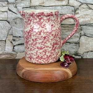 May include: A ceramic pitcher with a cream-colored base and a red speckled pattern. The pitcher has a curved handle and a spout for pouring. It sits on a round, wooden serving board. Decorative flowers are placed near the base of the pitcher.
