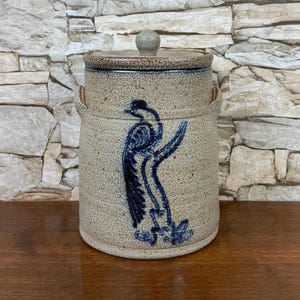 May include: A tan ceramic jar with a lid and a blue bird design. The jar has a round shape with two small handles on the sides. The lid has a small knob on top. The bird is painted in a stylized manner.