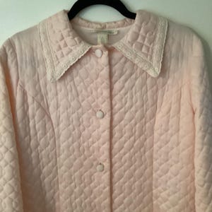 May include: A light pink quilted jacket with a diamond pattern, featuring a white lace-trimmed collar and button closure. The jacket has two pockets and is hanging on a black hanger.
