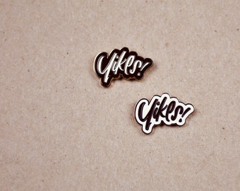 May include: Two enamel pins with the word "Yikes!" in a white, cursive font, outlined in black, and set against a gold border. The pins are on a tan background.