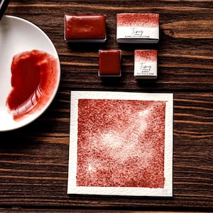 May include: A still-life composition featuring watercolor paints and supplies. The image showcases a palette with red paint, several rectangular paint blocks, and a watercolor painting. The paint blocks are labeled "Tiny Burnt Potter's Pink."
