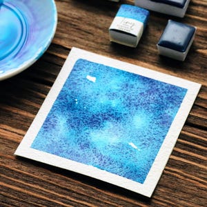 May include: A watercolour painting on white paper featuring a blue and turquoise galaxy design. Several small, rectangular watercolour paint pans and a ceramic dish with blue paint are arranged on a wooden surface.