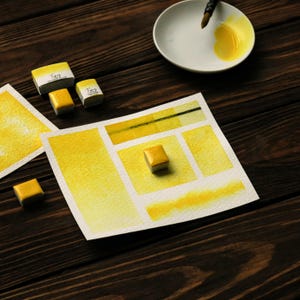 Op de afbeelding: Een stilleven van aquarelbenodigdheden. De scène omvat gele aquarelverf, een palet en een penseel. De verven zitten in kleine, vierkante napjes, sommige met het woord "Tiny" op de verpakking. Een aquarelkleurenkaart toont verschillende tinten geel.