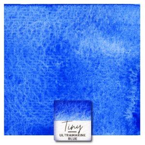 Op de afbeelding: Een vierkant staal van levendige ultramarijnblauwe aquarelverf met een getextureerde, korrelige wassing. Onderaan bevindt zich een klein wit label met de handgeschreven tekst Tiny en de gedrukte tekst ULTRAMARINE BLUE.