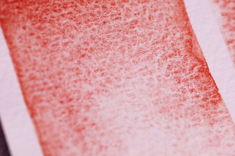 May include: Close-up of a red watercolour wash on paper. The wash is uneven and has a grainy texture. The paper is white and has a slight texture.