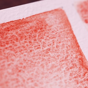May include: Close-up of a red watercolour wash on textured paper. The wash is uneven, with a lighter area at the top and a darker area at the bottom.