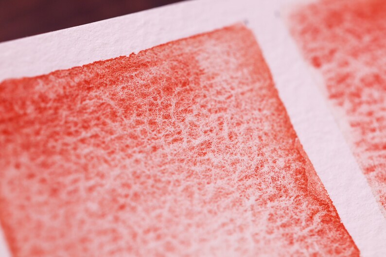 May include: Close-up of a red watercolour wash on textured paper. The wash is uneven, with a lighter area at the top and a darker area at the bottom.