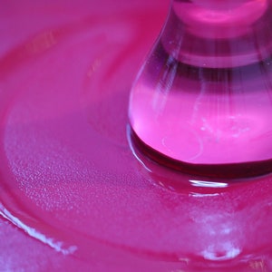Pu&ograve; includere: Primo piano di una bottiglia di vetro rosa con un liquido trasparente al suo interno. La bottiglia &egrave; appoggiata su una superficie rosa e ha creato un effetto increspato nel liquido.