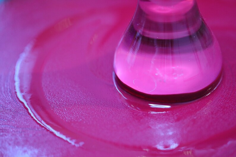 Pu&ograve; includere: Primo piano di una bottiglia di vetro rosa con un liquido trasparente al suo interno. La bottiglia &egrave; appoggiata su una superficie rosa e ha creato un effetto increspato nel liquido.