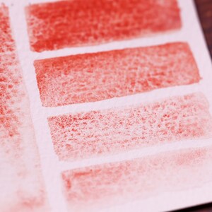 May include: A close-up of a white piece of paper with five red watercolour swatches. The swatches are arranged in a row, with the darkest swatch at the top and the lightest swatch at the bottom. The swatches are all slightly textured.