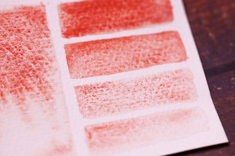 May include: A close-up of a white piece of paper with five red watercolour swatches. The swatches are arranged in a row, with the darkest swatch at the top and the lightest swatch at the bottom. The swatches are all slightly textured.