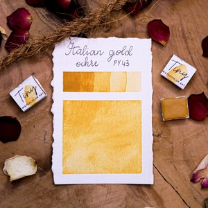 May include: Watercolor art supplies featuring a palette of Italian gold ochre (PY43) shades. The image shows a watercolor swatch, a small watercolor pan, and a Tiny Watercolor Art box. Rose petals and a twine accent the wooden surface.