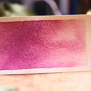 Op de afbeelding: Een aquarelverfstaal dat een verloop van roze naar paars laat zien, met de tekst "Ultramarine Pink".