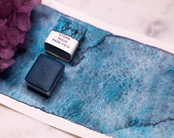 May include: Two watercolor paint pans, one is a dark blue color and the other is a light blue color with the text "STORM WAVE PB28, PB74" on the label. A blue watercolor wash is painted on a white paper.