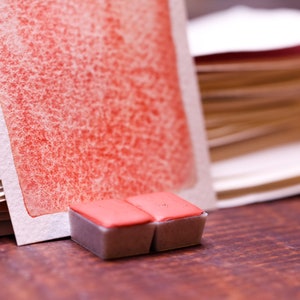 May include: Two small, rectangular watercolour paint pans with a red pigment. The pans are sitting on a wooden surface in front of a stack of watercolour paper.