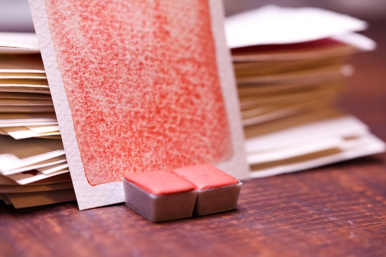 May include: Two small, rectangular watercolour paint pans with a red pigment. The pans are sitting on a wooden surface in front of a stack of watercolour paper.