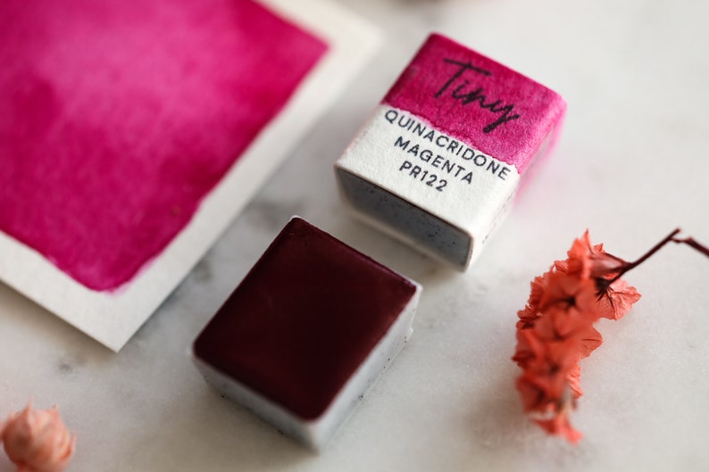 Pu&ograve; includere: Due pastiglie di acquerello, una aperta e una chiusa. La pastiglia aperta &egrave; di colore magenta vibrante e la pastiglia chiusa &egrave; di colore bordeaux scuro. La pastiglia chiusa ha il testo "Tiny Quinacridone Magenta PR122" sull'etichetta.