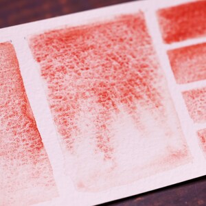 May include: A close-up of a white paper surface with red watercolour swatches. The swatches are arranged in a grid pattern, with some swatches being more saturated than others.