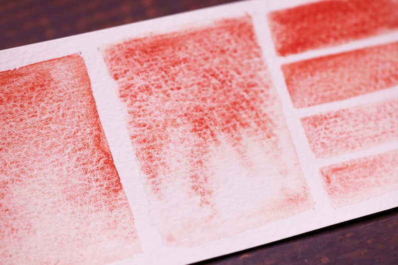 May include: A close-up of a white paper surface with red watercolour swatches. The swatches are arranged in a grid pattern, with some swatches being more saturated than others.