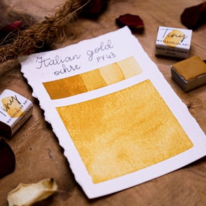 May include: Watercolor art set featuring "Italian gold ochre" paint. The image shows a swatch of the color, a small paint cube, and a label with the color name and code. The color is a warm, golden yellow.