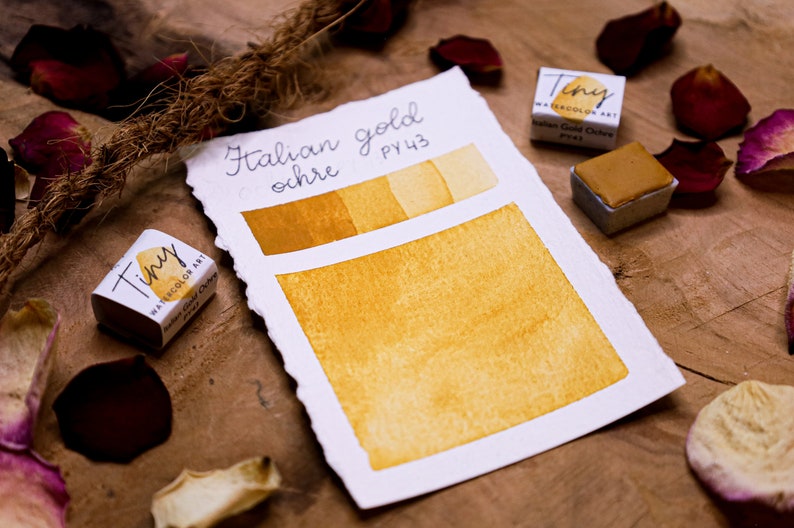 May include: Watercolor art set featuring "Italian gold ochre" paint. The image shows a swatch of the color, a small paint cube, and a label with the color name and code. The color is a warm, golden yellow.