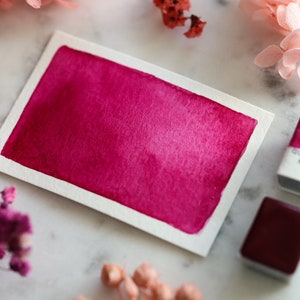 May include: A watercolor paint swatch in a vibrant magenta shade. The paint is applied to a white card and has a gradient effect. Two small pans of watercolor paint are also shown, one labeled "Tiny Quinacridone Magenta PR122".