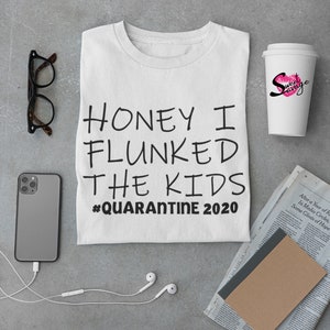 May include: White t-shirt with black text that reads "Honey I Flunked the Kids #Quarantine 2020". The t-shirt is laying flat on a gray surface with a pair of glasses, a smartphone, a pair of white earbuds, a brown notebook, a newspaper, and a white to-go cup with a pink lip design and the text "Sweet Savage" on it.