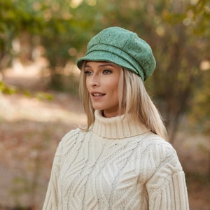 May include: A green newsboy cap with a textured herringbone pattern. The cap has a short brim and a rounded crown. The person is wearing a cream-coloured turtleneck sweater with a cable knit design. The background is a blurred view of a park.