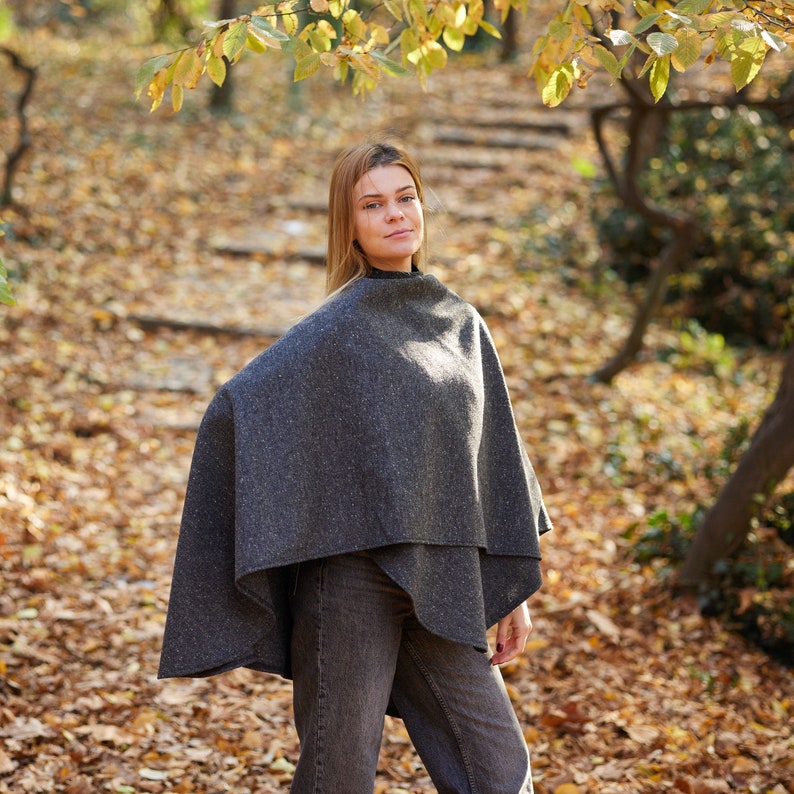 May include: A woman wearing a grey wool poncho in a woodland setting. The poncho is a simple, loose-fitting style with a wide neckline. The woman is standing on a path covered in fallen leaves.