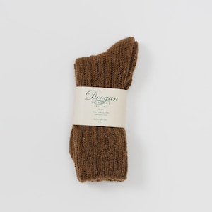 Könnte beinhalten: Ein Paar braune gerippte Wollsocken mit einem Papieretikett, das um die Mitte gewickelt ist. Auf dem Etikett steht "Doogan Donegal Ireland" und "Irish Walking Socks 100% pure wool". Die Socken haben einen warmen, erdigen Farbton.
