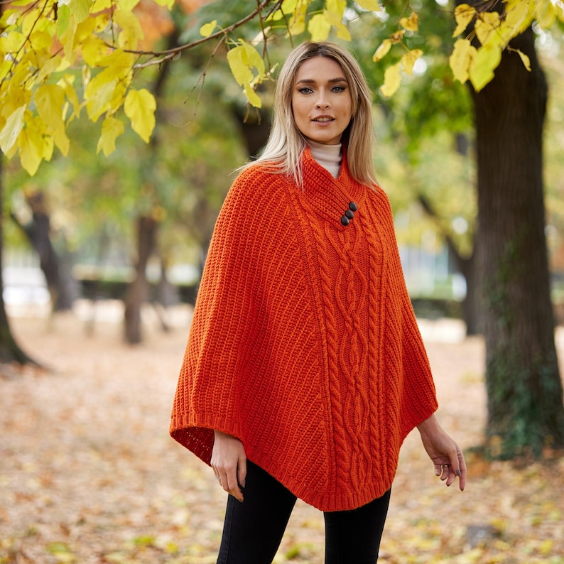 May include: A bright orange knitted poncho with a cable knit pattern and two brown buttons. The poncho is worn by a person standing in a park with fall foliage in the background.