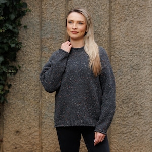 May include: A dark gray sweater with flecks of red, orange, and blue. The sweater has a crew neck and long sleeves. The person in the image is wearing black pants. The background is a textured stone wall.